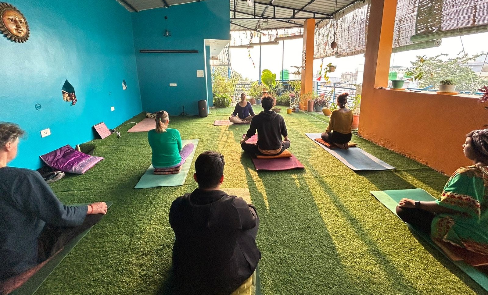 A yoga studio where Kaya teaches a group of foreigners. The walls are multi-colored, the floor is grass turf, and sunlight streams in through the open-air back wall.